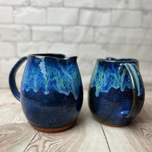 Load image into Gallery viewer, wheel thrown and handcrafted stoneware pottery pitcher in Blue world glaze. The pitcher is a dark cobalt blue on the bottom layer with a unique layering of turquoise and bright blue dripping down the top half of the pitcher. Two shown here, with the glaze dripping down the handle