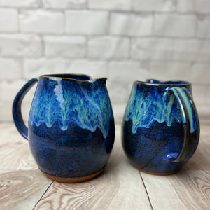 wheel thrown and handcrafted stoneware pottery pitcher in Blue world glaze. The pitcher is a dark cobalt blue on the bottom layer with a unique layering of turquoise and bright blue dripping down the top half of the pitcher. Two shown here, with the glaze dripping down the handle