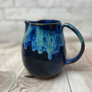 wheel thrown and handcrafted stoneware pottery pitcher in Blue world glaze. The pitcher is a dark cobalt blue on the bottom layer with a unique layering of  turquoise and bright blue dripping down the top half of the pitcher.
