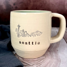 Load image into Gallery viewer,  Wheel thrown pottery city mug with the word &quot;seattle&quot; and an illustration of the seattle skyline inset on the outside. white outside, turquoise green glaze interior