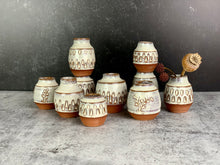 Load image into Gallery viewer, a collection of small bud vases with dried flowers in one of them. the vases are about 1.5-2 inches tall, wheelthrown in red stoneware, have hand carved facets and are glazed in white.