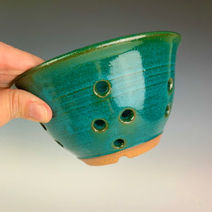 Berry colander in teal glaze on red clay. note the holes for drainage in the sides and the foot of the pot. shown being held by the artist for scale. holds a pint to a pint and a half