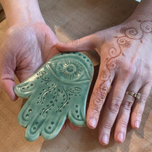 Load image into Gallery viewer, The artist holding a celadon green Hamsa Wall hanging which has been hand carved with a vine pattern. Her hands are also decorated in a vine pattern with Henna. wall decor amulet for protection. Fern Street Pottery