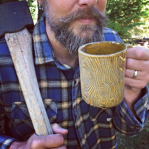 bearded woodsman with ax drinking out of a pottery mug that looks like a wooden mug or beer stein . Fern Street Pottery.