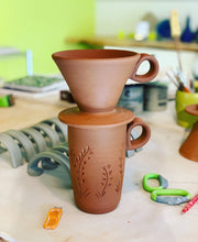 Load image into Gallery viewer, Travel mug and coffee pour over as they are being made in the pottery studio. The red clay is shown before it is glazed or fired. on the work table are handles for more mugs and pottery tools