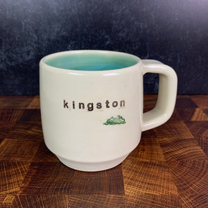 Wheel thrown pottery mug with "kingston" and an image of a ferry inset on the outside. white outside, turquoise green glaze interior