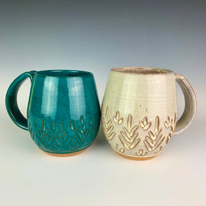 wheel thrown and hand carved mugs. the mug is thrown in red stoneware and glazed in Teal or white glaze at the edges of the mug. the mugs have delicate patterns carved into them. shown here in the fern/tree carved pattern