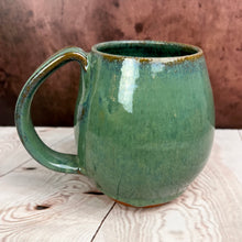 Load image into Gallery viewer, Northwest mug in Moss Green Glaze. This rich glossy glaze is  subtly mottled in shades of drippy green over a red stoneware clay.  The mug is shaped to keep your coffee warm longer.