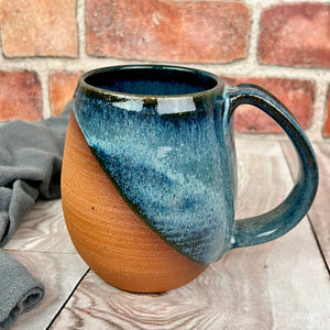 angle dipped mug, showing the beautiful red brown stoneware clay beneath the glaze. This mug is glazed in a dark blue glaze with swirls and drips of lighter mottled blue like a wave on the shore.wheel thrown pottery. Fern Street Pottery. Angle dipped mug.