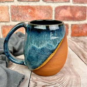 angle dipped mug, showing the beautiful red brown stoneware clay beneath the glaze. This mug is glazed in a dark blue glaze with swirls and drips of lighter mottled blue like a wave on the shore, with a crescent strip of light sand.wheel thrown pottery. Fern Street Pottery. Angle dipped mug.