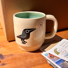 Load image into Gallery viewer, artisan made, wheel thrown pottery mug. porcelaina clay, with a cute crow or raven image that is painted on and carved into it. the crow speaks of love at it is speaking a heart in a speech bubble. 