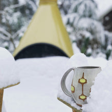 Load image into Gallery viewer, wheelthrown Pottery mug, hand glazed with MidMod pattern in white, orange and yellow, shown in a snow scene in front of a Mid Century style fireplace, in a snowy scene.