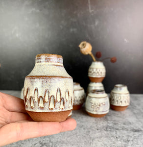 a collection of small bud vases , each one is hand carved with facets, with dried flowers in one of them. the vases are about 1.5-2 inches tall, wheelthrown in red stoneware, 