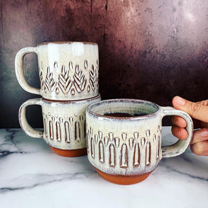 A collection of wheelthrown carved mugs glazed in rustic white glaze. the carved facets show through the white glaze. these mugs are stackable and dishwasher safe too.