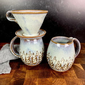 wheel thrown and hand carved mugs. the mug is thrownin red stoneware which shows throught the white glaze at the edges of the mug. the mugs have delicate patterns carved into them. shown here in the fern/tree carved pattern, with a matching coffee pour over