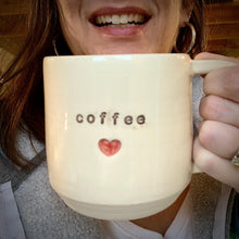 Load image into Gallery viewer, The artist holding a Coffee Love Mug, with the word coffee stamped into the mug, a red heart is stamped in and painted below it.. White clay, turquoise glaze on the inside. this mug was wheel thrown and hand stamped and colored at fern street pottery.