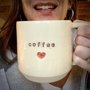 The artist holding a Coffee Love Mug, with the word coffee stamped into the mug, a red heart is stamped in and painted below it.. White clay, turquoise glaze on the inside. this mug was wheel thrown and hand stamped and colored at fern street pottery.