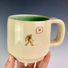 Load image into Gallery viewer, Sasquatch with a heart bubble on an artist made, wheel thrown pottery mug. Beautiful porcelaina creamy white clay with a turquoise glaze on the inside.