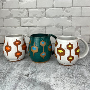 wheelthrown Pottery mug, hand glazed with MidMod pattern, shown here in white and orange, teal and turquoise, white with yellow. each with the deep red clay showing in the resist pattern. Fern Street Pottery