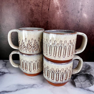 A Stacking collection of wheelthrown carved mugs glazed in rustic white glaze. the carved facets show through the white glaze. 