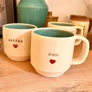 Mom love mug and a coffee love mug, each with a heart stamped into the mug under the text. White clay, turquoise glaze on the inside. this mug was wheel thrown and hand stamped and colored at fern street pottery.