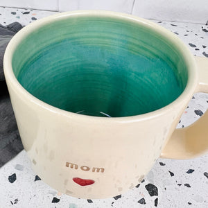 Mom love mug, with mom and a heart stamped into the mug. White clay, turquoise glaze on the inside. this mug was wheel thrown and hand stamped and colored at fern street pottery.