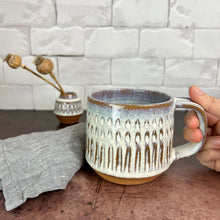 Load image into Gallery viewer, A wheelthrown carved mug glazed in rustic white glaze. the carved facets show through the white glaze. two matching vases are in the background.