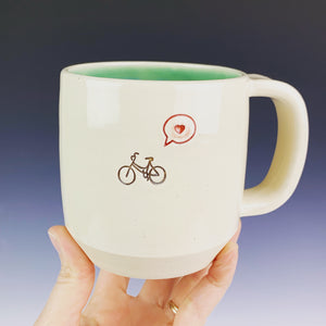 Bike love mug for the bicycle lover. white cylindrical mug with a pug stamped in in brown, and a heart in a speech bubble in red. Turquoise green interior on this mug.