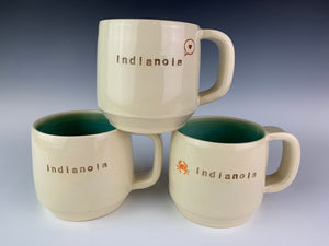 stack of three indianola mugs. White cylidrical mugs, thrown on the potters wheel. glazed turquoise on the inside. each one is stamped with the word "indianola" and another image