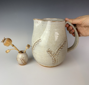 Carved pitcher in speckled white. the red clay and speckled white glaze work well together to show the carved detail. the carved pattern on the vase is reminiscent of vines or growing plants. shown here with a carved bud va