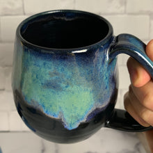 Load image into Gallery viewer, Aurora Borealis mug, blue over black glaze, northwest style coffee mug thrown pottery, with large pulled handle. shown held by the artist