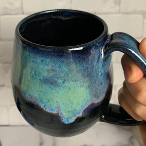 Aurora Borealis mug, blue over black glaze, northwest style coffee mug thrown pottery, with large pulled handle. shown held by the artist