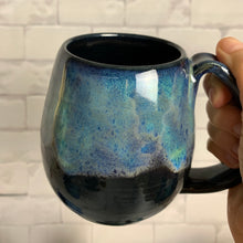 Load image into Gallery viewer, Aurora Borealis mug, blue over black glaze, northwest style coffee mug thrown pottery, with large pulled handle. shown held by the artist