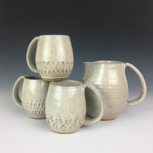 Load image into Gallery viewer, wheel thrown and hand carved mugs. the mug is thrownin red stoneware which shows throught the white glaze at the edges of the mug. the mugs have delicate patterns carved into them. shown here with a matching pitcher