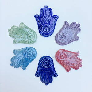 An array of Hamsas in a variety of colors, each one hand carved with an eye and a vine pattern, or a fish pattern. Cobalt Blue, Rose Pink, Lavender Purple, Celadon Green, Skye Blue.