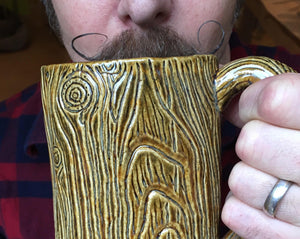 lumberjack style, handlebar mustache, man drinking out of a large pottery mug that is carved to look like tree bark. Fern Street Pottery.