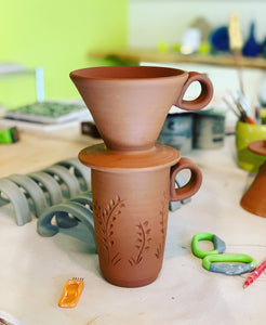 Travel mug and coffee pour over as they are being made in the pottery studio. The red clay is shown before it is glazed or fired. on the work table are handles for more mugs and pottery tools