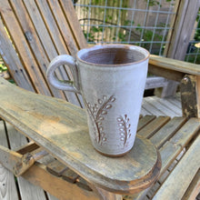 Load image into Gallery viewer, tall latte size travel mug with finger loop handle. vine carvings and white glaze. shown on adirondak chair