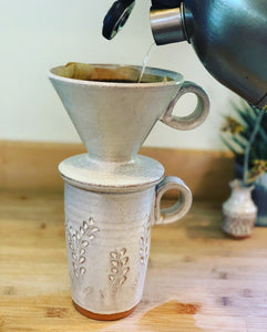 travel mug shown with coffee pour over. both are thrown on the potters wheel in red clay, the mug is carved with a vine pattern and they are glazed in speckled white glaze