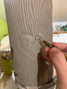 woodgrain textured vase, cylindrical in shape with heart and initials carved into texture. the artist carving a vase that is reminiscent of a tree with carved initials