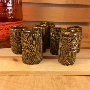 a collection of 7 lumberjack shot glasses shown with bourbon. pottery carved to look like wood
