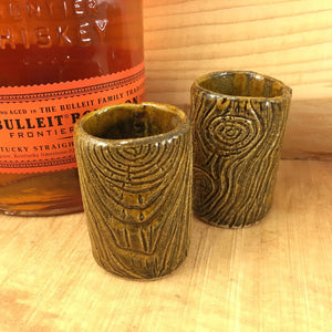 two lumberjack shot glasses, pottery carved to look like woodgrain, shown with a bottle of bourbon
