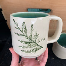 Load image into Gallery viewer, a custom handcrafted mug with a Cedar branch impressed into the side. the interior is glazed in a matching green