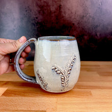 Load image into Gallery viewer, a wheel thrown and hand carved mug is held by the artists. the mug is thrownin red stoneware which shows throught the white glaze at the edges of the mug. the mug has a delicate vine pattern carved into it.