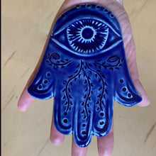 Load image into Gallery viewer, The artist holding a hand carved ceramic Hamsa in cobalt blue. The hamsa has an eye and a vine pattern carved into it.