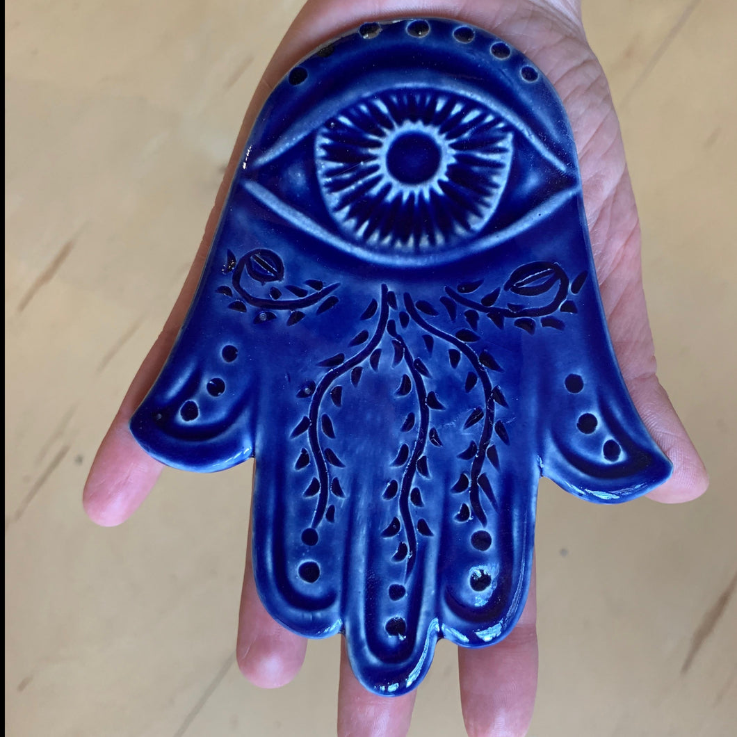 The artist holding a hand carved ceramic Hamsa in cobalt blue. The hamsa has an eye and a vine pattern carved into it.