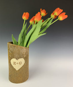 A pottery vase, with woodgrain texture and initials and a heart carved into the tree-like surface. shown with tulips