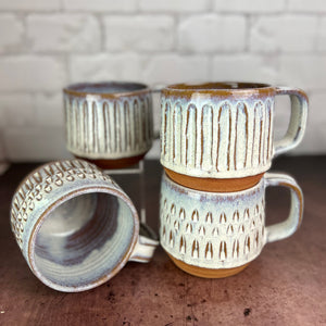 a collection of wheelthrown, carved mugs glazed in rustic white glaze. the carved facets show through the white glaze. 