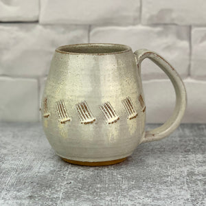 a wheel thrown and hand carved mug sits in a kitchen setting. the mug is thrownin red stoneware which shows throught the white glaze at the edges of the mug. the mug has a repeated line pattern scratched pattern carved into it.